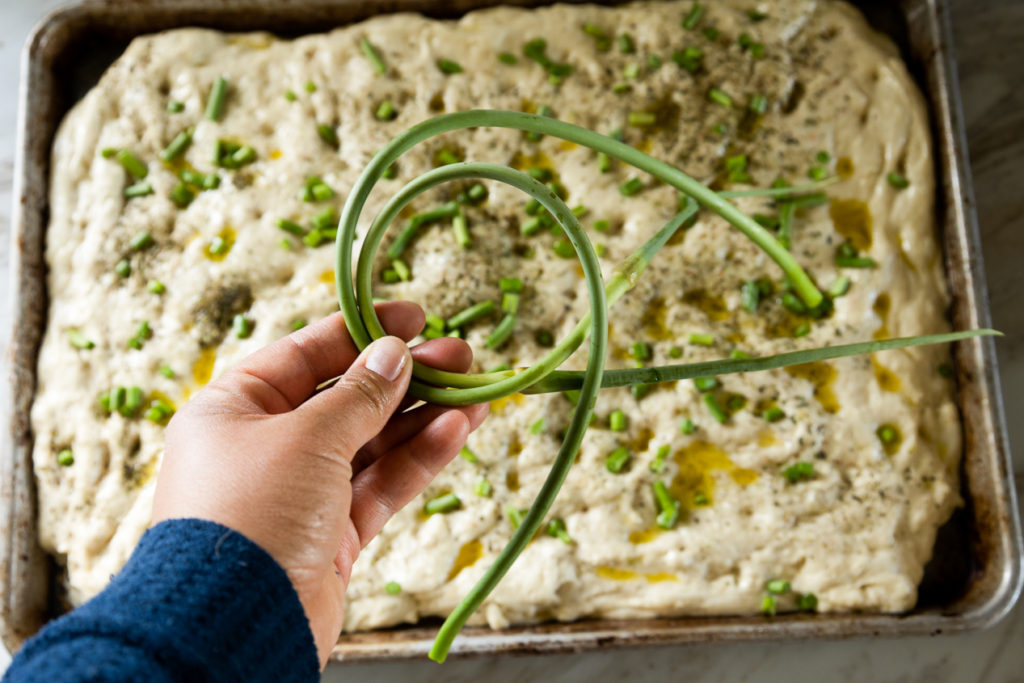 hand holding garlic scapes over tray of focaccia bread 